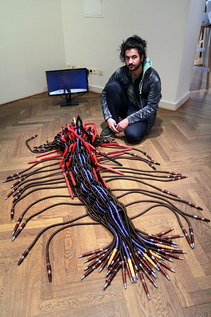 Der Künstler Dachil Sado hockt auf dem Boden der Galerie vor einem Bildschirm und einem Objekten aus Shisha-Schläuchen.