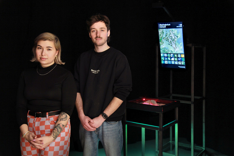Die zwei Künstlerinnen stehen in einem dunklen Raum vor einem schwarzen Gestellt mit einem Monitor auf dem eine Insel angezeigt wird.