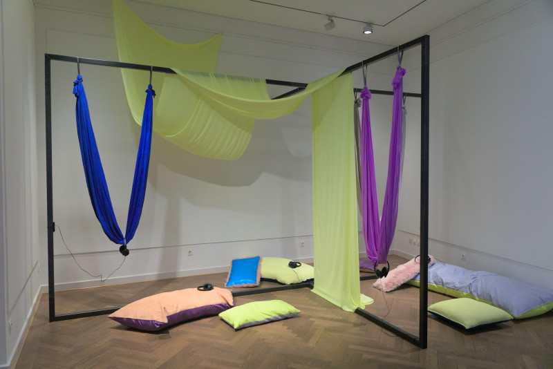 Das Bild zeigt eine raumfüllende Stahlkonstruktion mit farbigen Textilien und Sitzmöglichkeiten und Kissen.