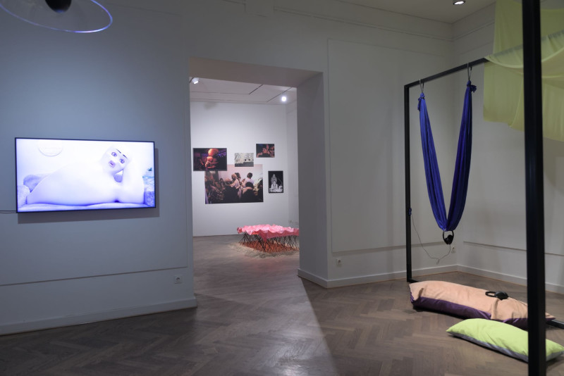 Das Bild zeigt einen Bildschirm mit Videoarbeit und daneben Stahlrahmen mit farbigen Textilien