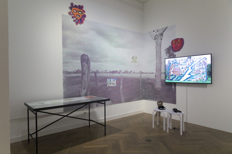In front of a photo wallpaper hangs a screen with a video work, next to it is a table with images and texts
