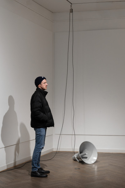 Man sieht einen grauen Industrielautsprecher auf dem Boden liegen, ein schwarzes Kabel führt zur Decke und wieder um Boden. Daneben steht ein Mann und guckt nach rechts.