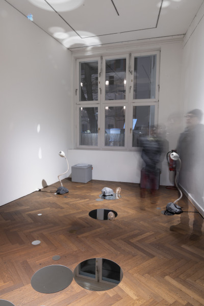 Man sieht einen Galerieraum mit runden Spiegel und ein paar Steinen auf dem Boden. Zwei weiße kleine Roboterstängel stehen im Raum. Menschen bewegen sich verschwommen durch den Raum.