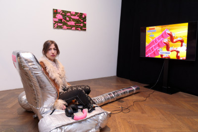 Woman in furry jacket sits in gamingchair playing video games on large screen in gallery space with pink artwork on wall