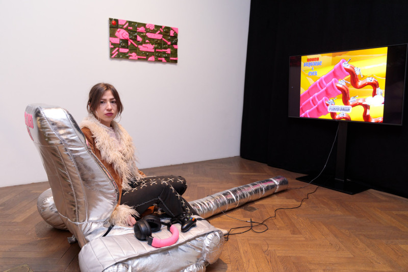 text: >
  Künstlerin sitzt auf silbernem
  Gamingstuhl in Panzerform vor Fernseher
  mit Videospiel in einer
  Galerieausstellung
decorative: false
source: reviewed