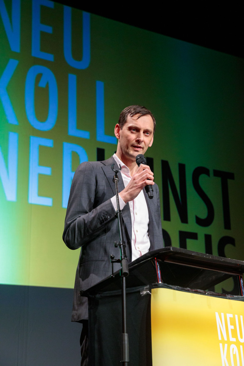 Borough Mayor Martin Hikel stands at a lectern with a microphone in his hand. The logo of the Neukölln Art Prize 2026 is projected in the background.