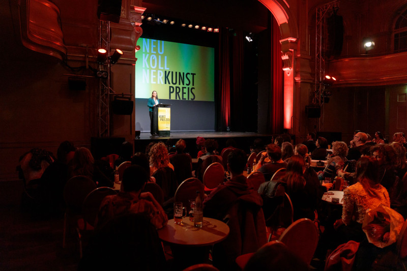 Menschen sitzen bei roter Beleuchtung in seinem Saal. Auf der Bühne spricht Janine Wolter, Bezirksstadträtin für Bildung, Kultur und Sport.