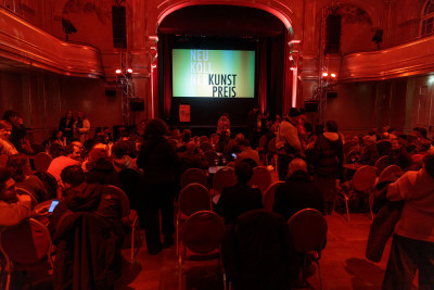 A hall lit in red with a stage at the far end. People fill the hall.