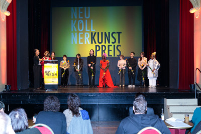 The nominees for the art prize, the curator, the gallery director, and the department head are standing on a stage. In front of them, several people are sitting on chairs facing the stage.