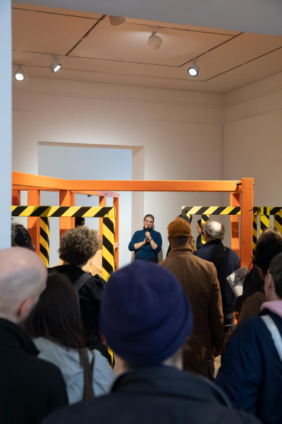 Artist speaking to audience at gallery opening with orange construction barrier installation in foreground