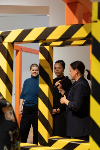 Three women stand in an art installation featuring yellow and black striped barriers, with one person holding a microphone