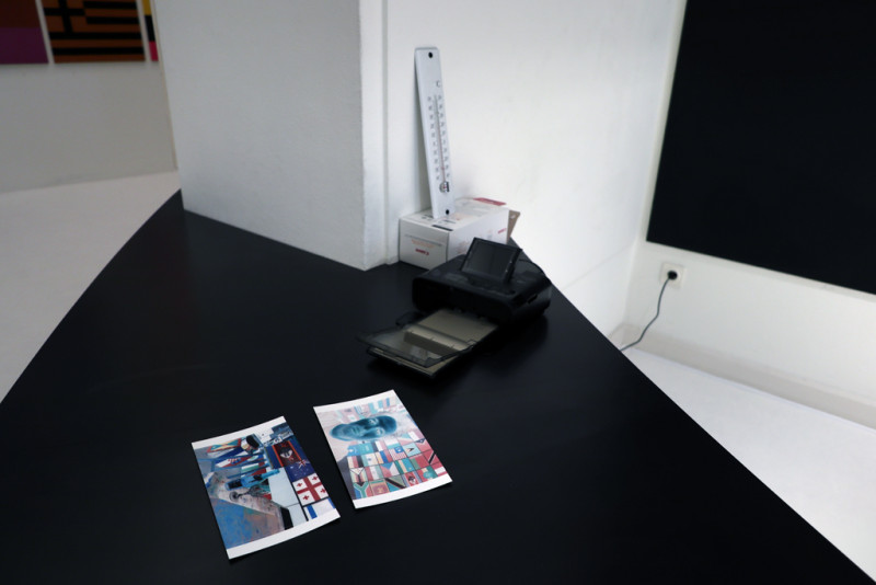 Auf einem schwarzen Tisch steht ein kleiner Drucker und liegen zwei invertierte Fotografien.