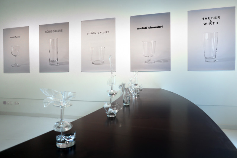 Auf einem schwarzen Tisch, der einen leichten Bogen im Raum macht, stehen aus Glasbruchstücken neu zusammengesetzte Objekte. Dahinter an der Wand hängen 5 Prints auf denen jeweils ein Glas und ein bekannter Kunstgalerienname abgebildet ist.