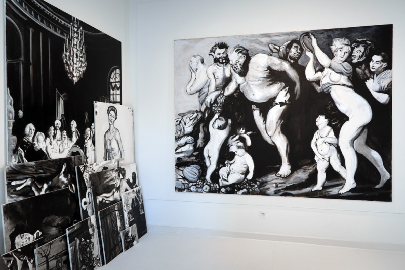 Mehrere verschiedenformatige schwarz-weiße Bilder stehen auf dem Boden an die Wand gelehnt. Ein großformatiges, altmeisterlich auschauendes, schwarz-weißes Gemälde hängt daneben an der Wand.
