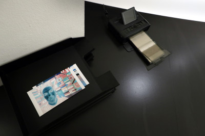 Auf einem schwarzen Tisch steht ein kleiner Drucker und liegen zwei invertierte Fotografien.