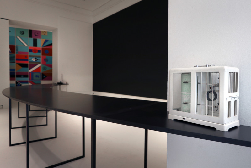 Ausstellungsansicht mit einem langen schwarzen Tisch, der sich durch mehrere Galerieräume zieht. Im Vordergrund steht ein Klimaüberwachungsgerät, wie man es aus Museen kennt.
