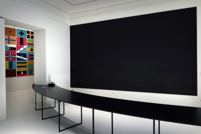 Ausstellungsansicht mit einem langen schwarzen Tisch, der sich durch mehrere Galerieräume zieht. Im Vordergrund steht ein Klimaüberwachungsgerät, wie man es aus Museen kennt.