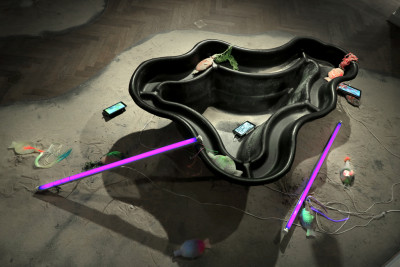Two neon tubes with black light lie on a pile of sand next to a garden pond bowl and several Ipads with video works