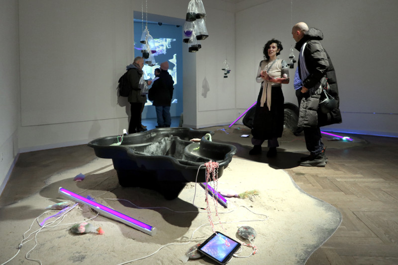 Im Vordergrund eine Gartenteichwanne auf Sand mit verschiedenen Elementen der Installation, im Hintergrund sind Personen zu sehen.