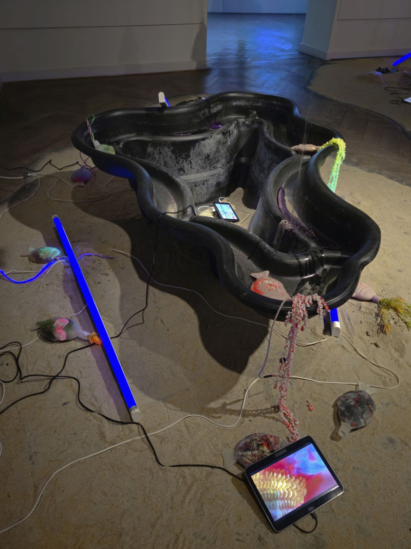 Zwei Neonröhren mit Schwarzlicht liegen auf einem Sandhaufen neben einem Gartenteich-Schale und mehreren Ipads mit Videoarbeiten