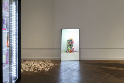View of a refrigerator containing frozen objects in neon colours and, to the right, a monitor showing a video work and stickers on the floor.