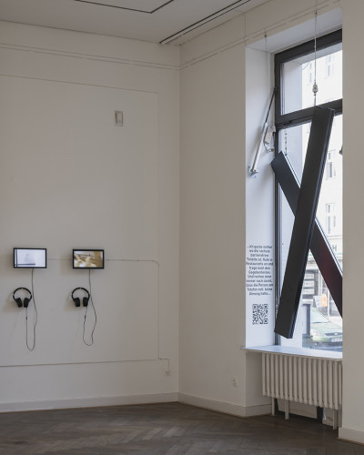 On the left side of the picture, two screens hang on the wall; on the right, you can see the window with two neon signs forming a cross and a wall text.