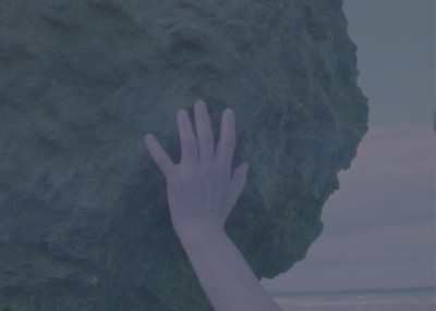 Eine Hand greift von unten gegen einen Felsbrocken. Im Hintergrund ist das Meer und der Himmel zu sehen.