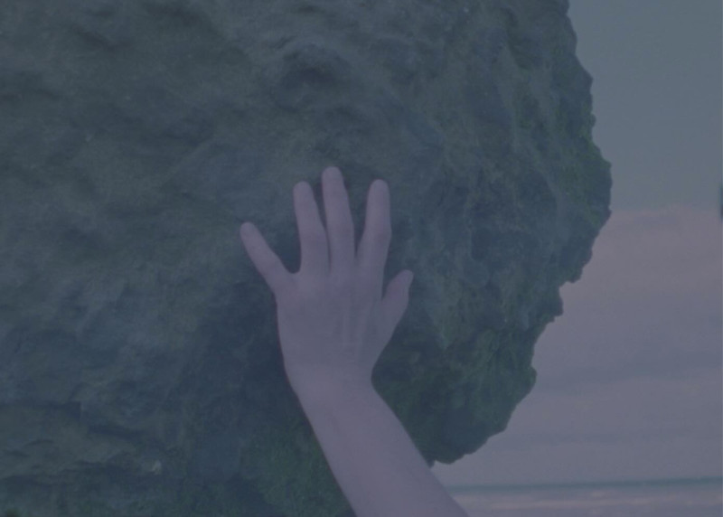 Eine Hand greift von unten gegen einen Felsbrocken. Im Hintergrund ist das Meer und der Himmel zu sehen.