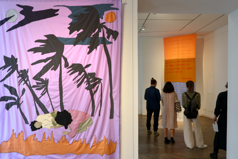 Stoffbild mit Palmen und zwei Frauen im Vordergrund, im Hintergrund Besucher:innen der Ausstellung vor einem Stoffbanner