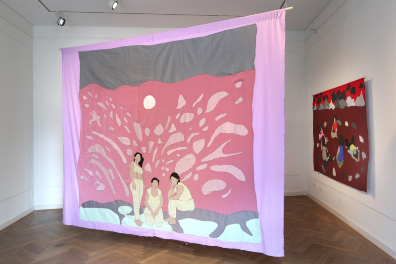 großes Stoffbild das in der Mitte des Raums von der Decke hängt und drei Frauen zeigt