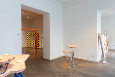 Ausstellungsraum mit Holztischen, Metallstuhl und rot-gelber Absperrung in einem angrenzenden Raum