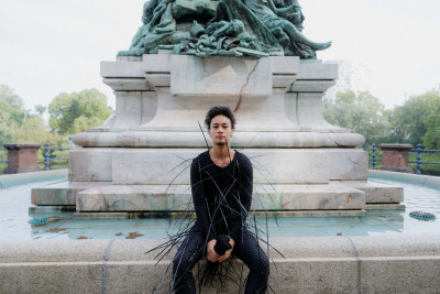 The artist in black clothing with spikes is sitting in front of a fountain.