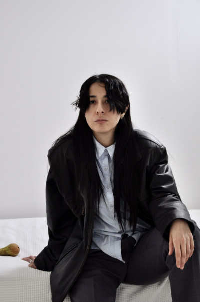 Die Künstlerin sitzt im rechten Bildvordergrdund auf einer weißen Decke, im Hintergrund ist eine weiße Wand zu sehen.