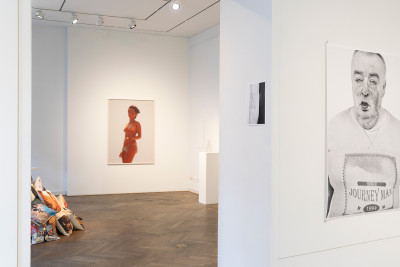 Galerieraum mit Kunstwerken an den Wänden, darunter eine Fotografie einer halbnackten Frau und ein Porträt eines Mannes, sowie Kunstobjekte auf dem Boden