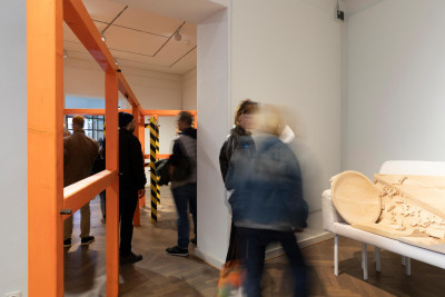 Exhibition visitors in a gallery space. Wooden sculptures are placed on a sofa, and a large wooden structure stands in the room