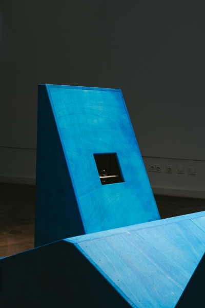 Blaue geometrische Holzinstallation mit schwarzem rechteckigen Fenster, installiert in einem Galerieraum mit Holzboden und weißen Wänden