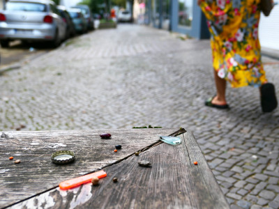Oben rechts im Bild läuft eine Person, deren Beine nur zu sehen sind, auf dem Fußweg - links unten ist ein Holzbank mit kleinen Gegenständen daruf