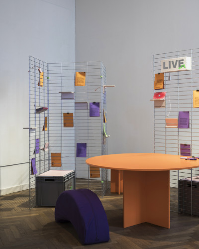 Eine Bibliotek mit einem orngefarbenen Tisch, einer violetfarbenen Sitzbanane und Kurztexten