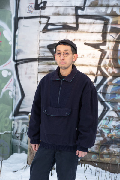 Der Künstler steht vor einer mit Graffiti übersäten Wand und schaut in die Kamera.