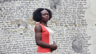 Eine Frau in einem rot weißen Kleid mit roten Lippen und Afro-Frisur steht vor einer beschriebenen Wand