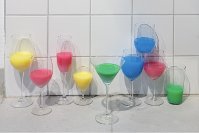 Arrangement aus neun Gläsern mit farbigen Flüssigkeiten in Gelb, Rosa, Blau und Grün vor weißer Fliesenwand